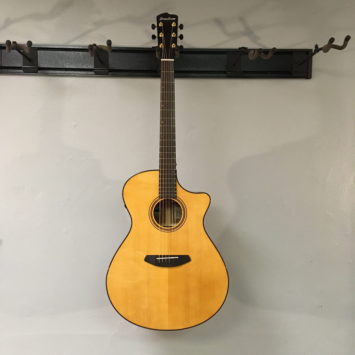 A Breedlove Performer Pro Concerto Aged Toner acoustic guitar with a natural wood finish hangs on a wall-mounted rack against a light gray background.