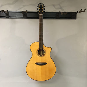 A Breedlove Performer Pro Concerto Aged Toner acoustic guitar with a natural wood finish hangs on a wall-mounted rack against a light gray background.