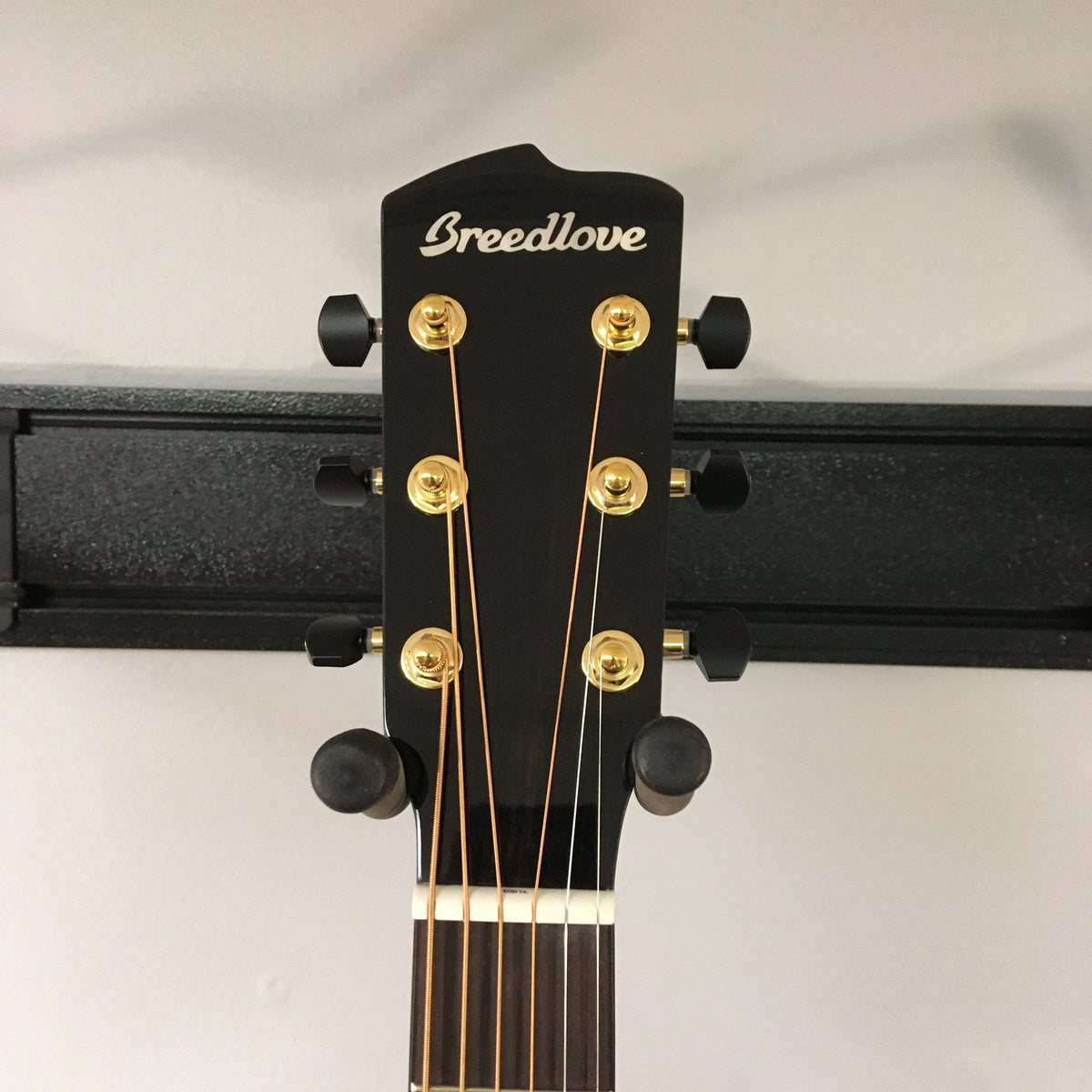 Close-up of the headstock of a Breedlove Performer Pro Concerto Aged Toner w/Gator Gigbag, featuring gold tuning pegs on a wall hanger against a light background, highlighting its signature refined acoustic tone.