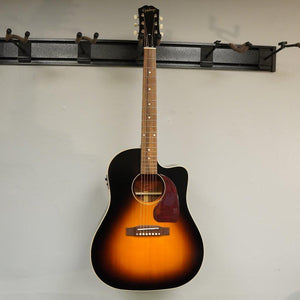 Epiphone Masterbilt J-45 EC Aged Vintage Sunburst Gloss guitar leaning against a wall, showcasing its classic sloped shoulder body and intricate detailing.