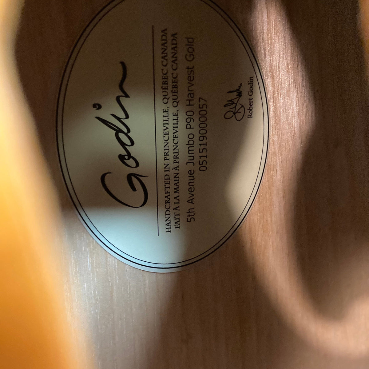 Close-up of Godin 5th Avenue Jumbo P90 Harvest Gold label on a table, highlighting text details and part of the guitar’s headstock.