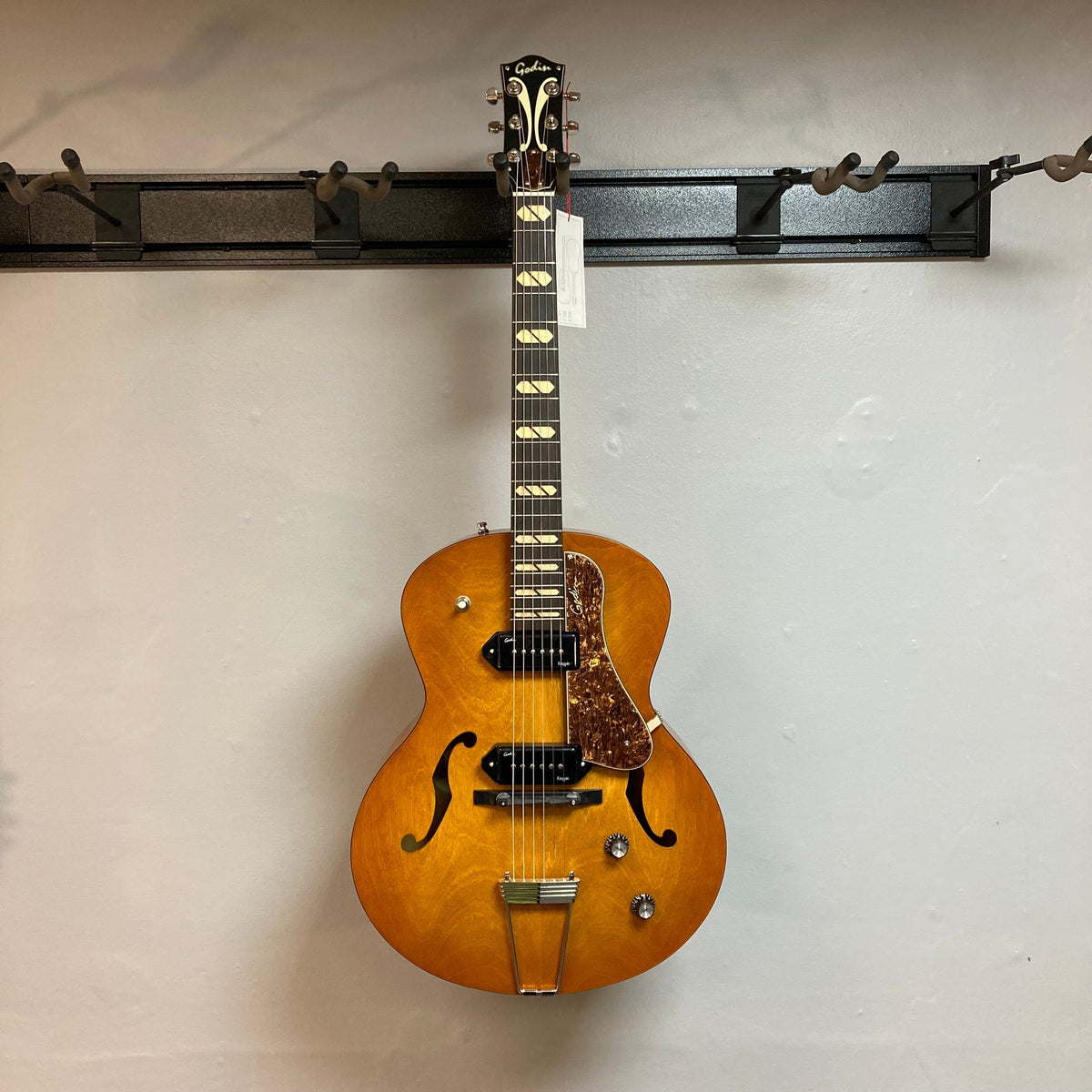 Godin 5th Avenue Jumbo P90 Harvest Gold guitar with redesigned headstock and large fret markers, displayed on a wall.