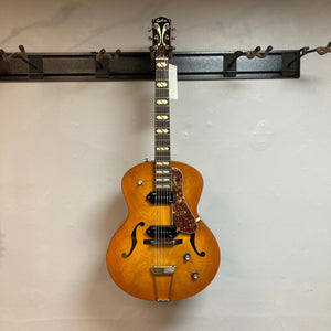 Godin 5th Avenue Jumbo P90 Harvest Gold guitar with redesigned headstock and large fret markers, displayed on a wall.