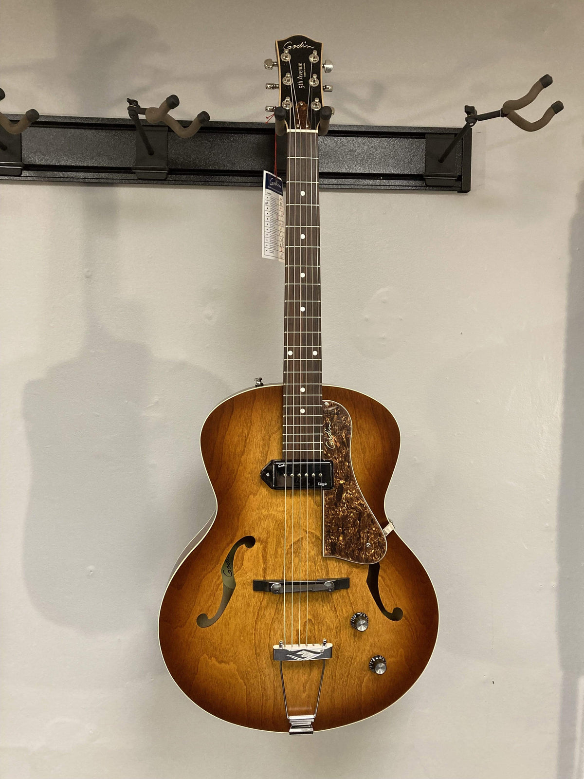 Godin 5th Avenue Kingpin P90 Cognac Burst guitar with archtop, floating pickguard, and high-gloss headstock, mounted on a wall.