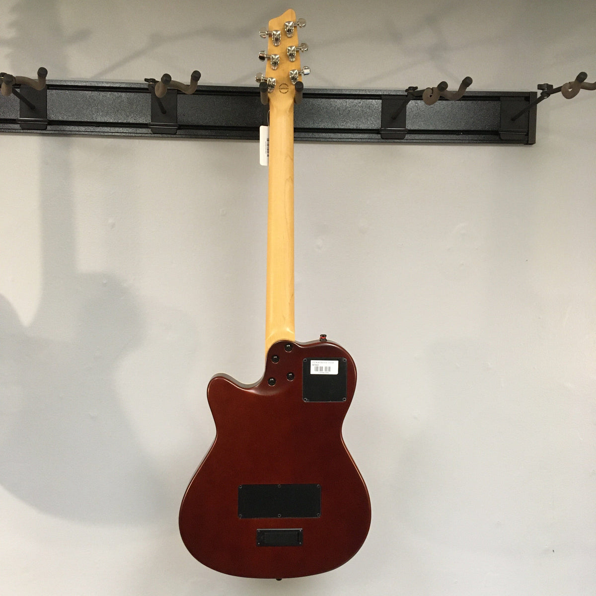 Godin A6 Ultra Baritone Burnt Umber guitar hanging on a wall, showcasing its chambered body and custom mini-humbucker pickup.
