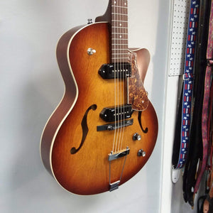 Godin 5th Avenue CW Kingpin II Cognac Burst electric guitar with black knobs and dual P90 pickups, displayed on a wall.