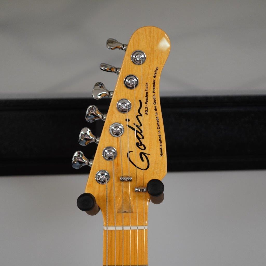 Close-up of a Godin RG3 Passion Lace Sensor Hot Gold Indigo Blue electric guitar headstock with six tuners and the Godin logo. The background is blurred with a black horizontal line. Includes gig bag.