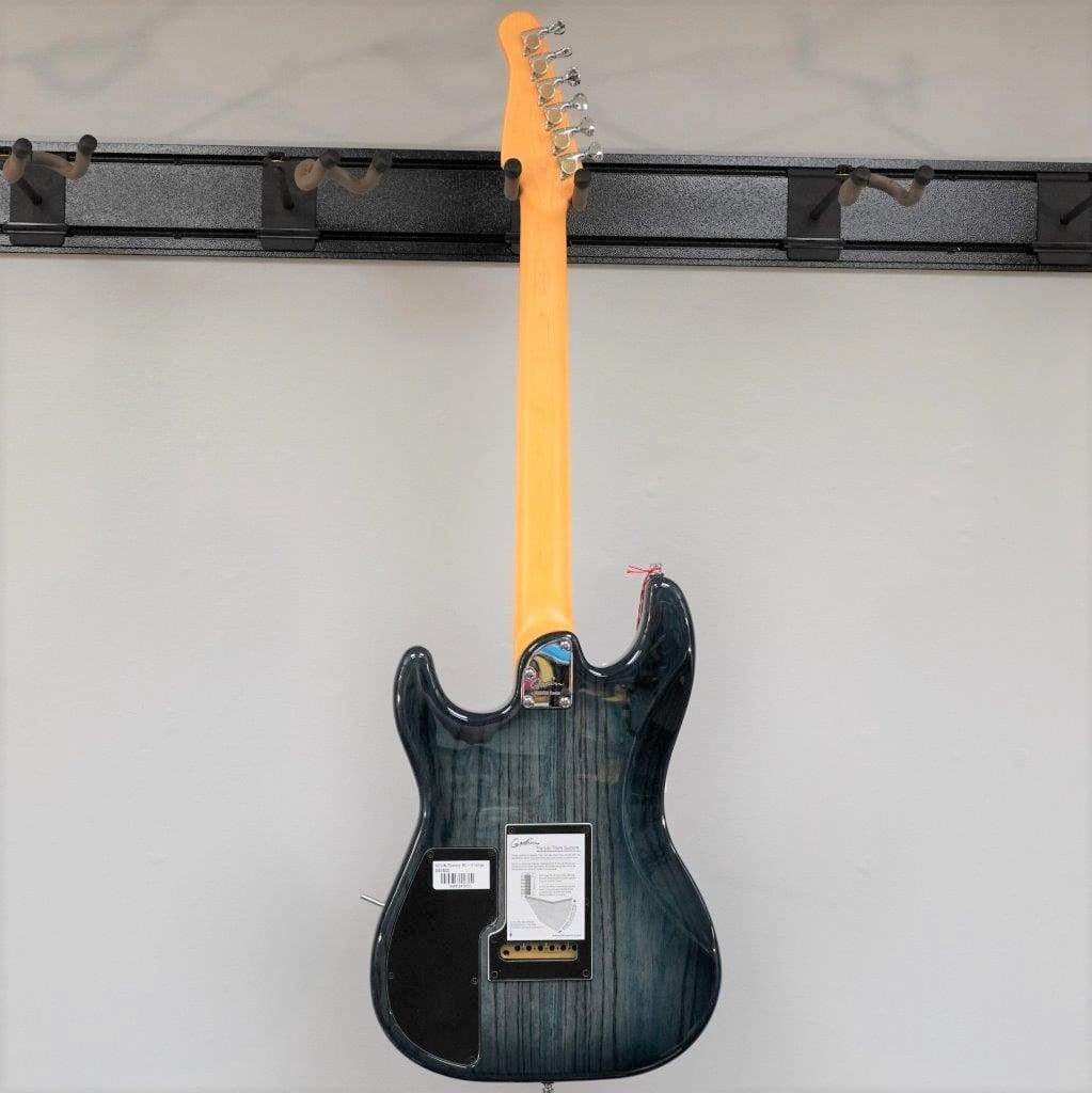 A Godin RG3 Passion Lace Sensor Hot Gold electric guitar in Indigo Blue with a gig bag is displayed on a wall rack, shown from the back. The light wood neck, tags, and barcode label are visible.