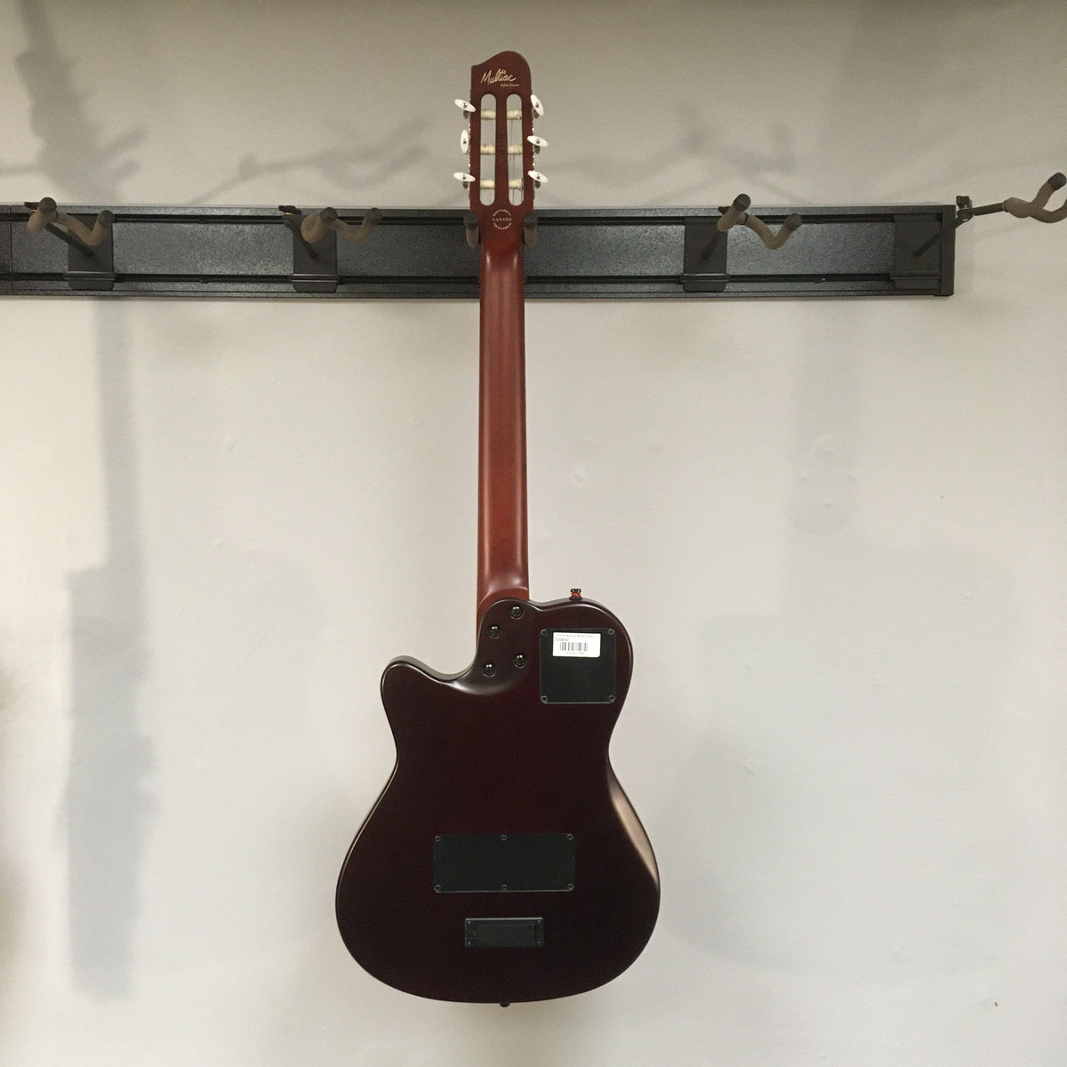 Godin Multiac Nylon Encore SG SF guitar with visible finish blemishes, mounted on a wall, showcasing its solid cedar top and dual source electronics.