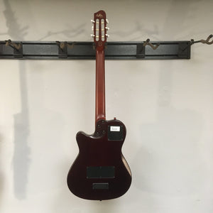 Godin Multiac Nylon Encore SG SF guitar with visible finish blemishes, mounted on a wall, showcasing its solid cedar top and dual source electronics.