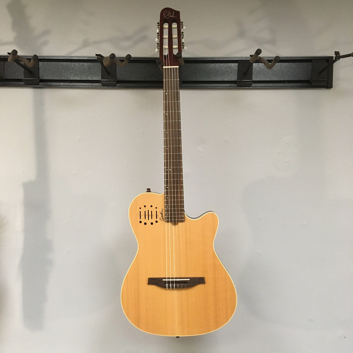 Godin Multiac Nylon Encore SG SF guitar with visible finish blemishes, mounted on a wall. Close-up shows dual-source electronics and blend controls. Comes with gig bag.