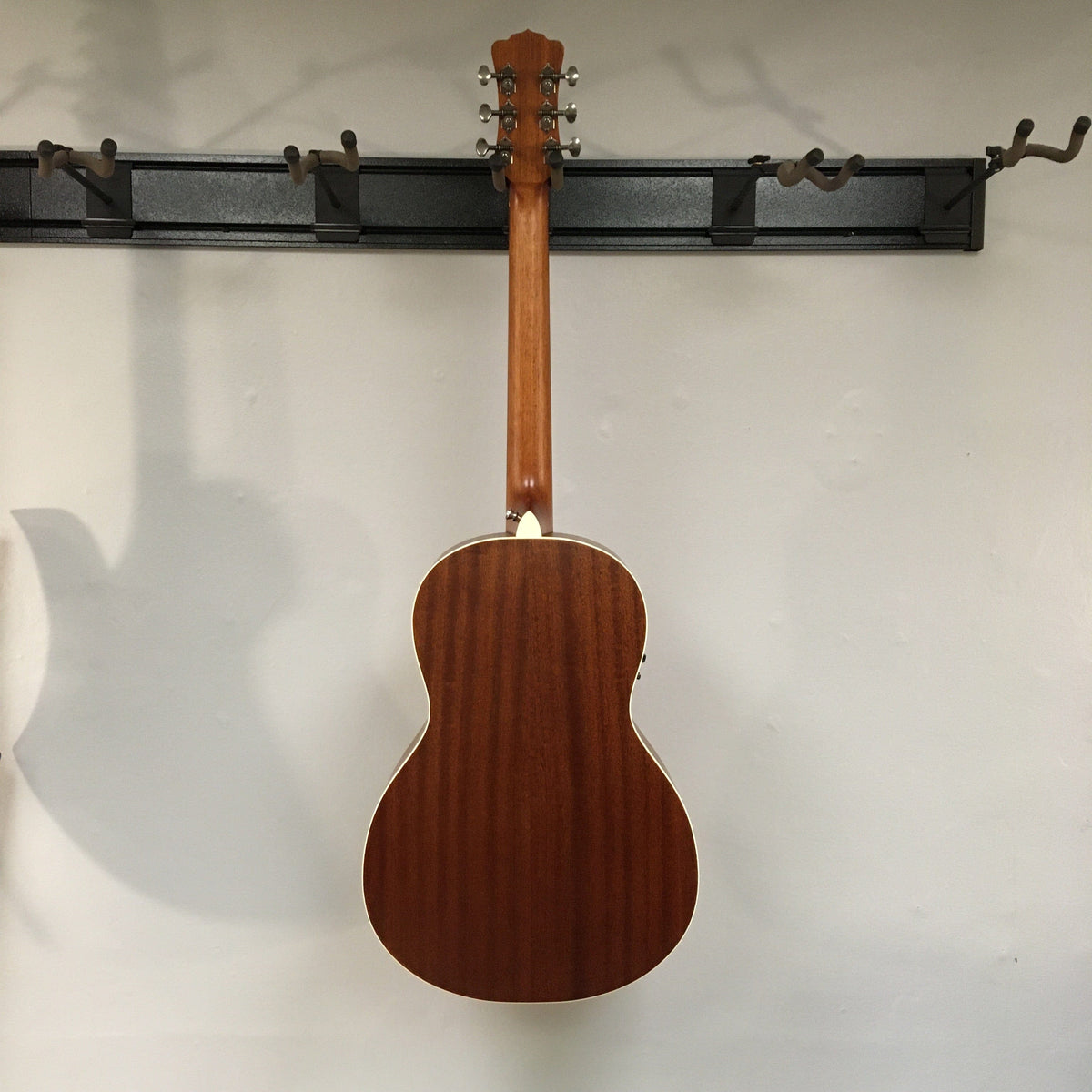 Luna Art Vintage Parlor Solid Top Acoustic-Electric guitar hanging on a wall, showcasing its distressed vintage brownburst finish and open gear tuners.