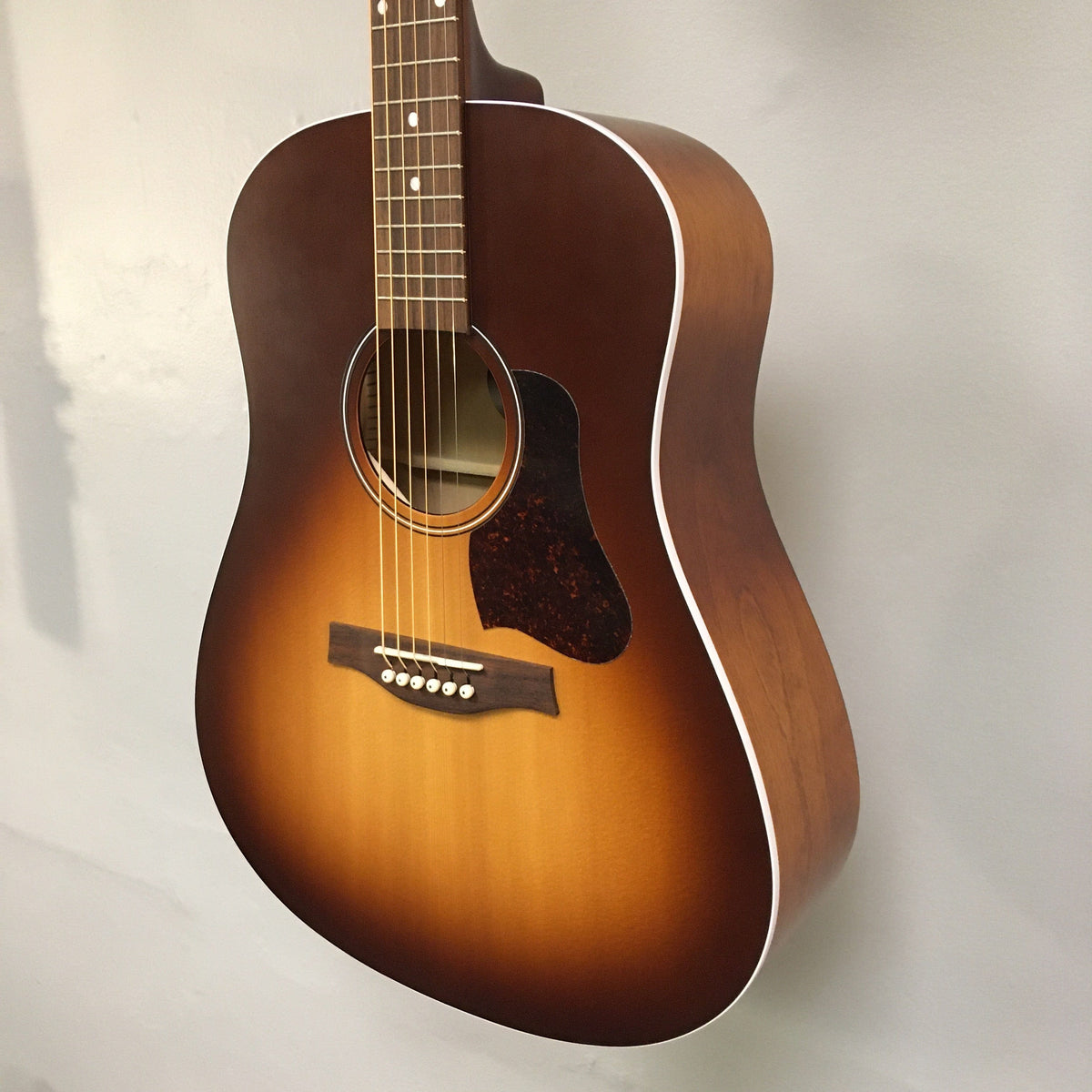 A Seagull Entourage Autumn Burst Acoustic Guitar by Seagull hangs on a light wall, featuring six strings, a tortoiseshell pickguard, and white binding along the body edges.