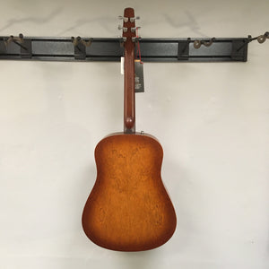 A Seagull Entourage Autumn Burst Acoustic Guitar hangs on a wall-mounted holder, back facing out to reveal its smooth wooden finish and chrome tuning pegs against a white wall.