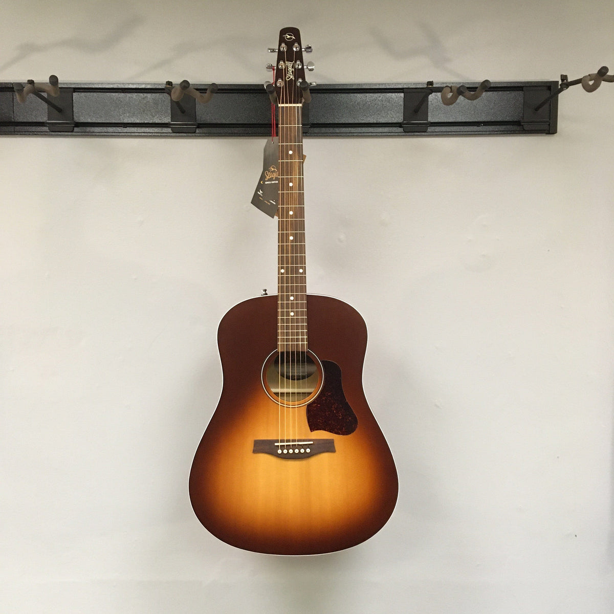 A Seagull Entourage Autumn Burst Acoustic Guitar by Seagull, featuring six strings, hangs on a wall-mounted rack against a plain white wall with a tag attached to its headstock.