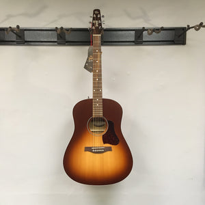 A Seagull Entourage Autumn Burst Acoustic Guitar by Seagull, featuring six strings, hangs on a wall-mounted rack against a plain white wall with a tag attached to its headstock.