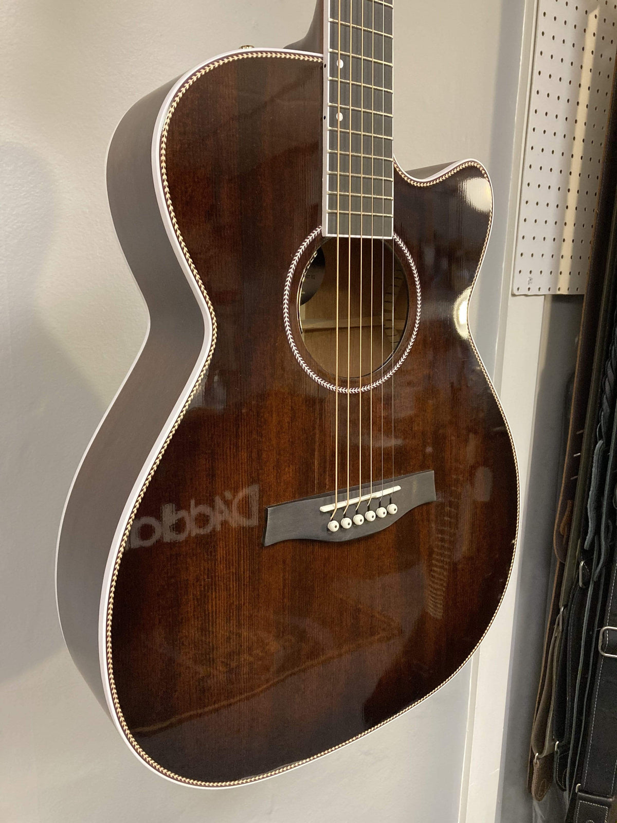 A Seagull Artist Mosaic CH CW Acoustic-Electric Guitar - Bourbon Burst (with TRIC Case) hangs on a wall, showing its cutaway body, white binding, six strings, and pearl inlays near a pegboard.