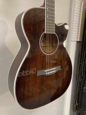 A Seagull Artist Mosaic CH CW Acoustic-Electric Guitar - Bourbon Burst (with TRIC Case) hangs on a wall, showing its cutaway body, white binding, six strings, and pearl inlays near a pegboard.