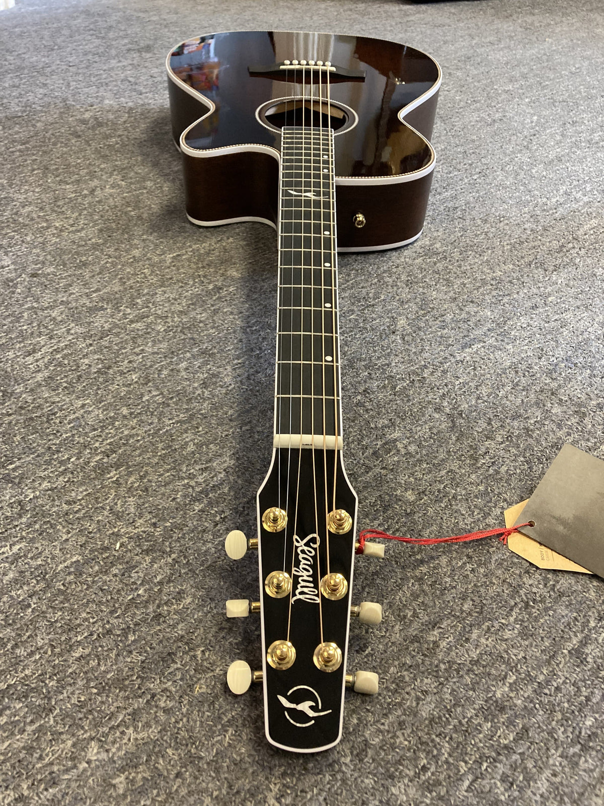 A Seagull Artist Mosaic CH CW Acoustic-Electric Guitar - Bourbon Burst (with TRIC Case) with a dark wood body and light trim rests on a gray carpet, headstock with brand name and tuning pegs closest to the camera, tag on one peg.