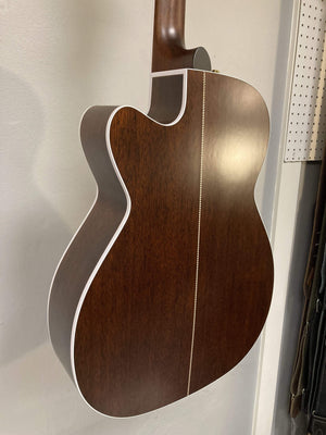 The image shows the back of a Seagull Artist Mosaic CH CW Acoustic-Electric Guitar - Bourbon Burst, featuring a smooth dark wood finish, white edge binding, and a decorative center inlay. The guitar is upright against a wall.
