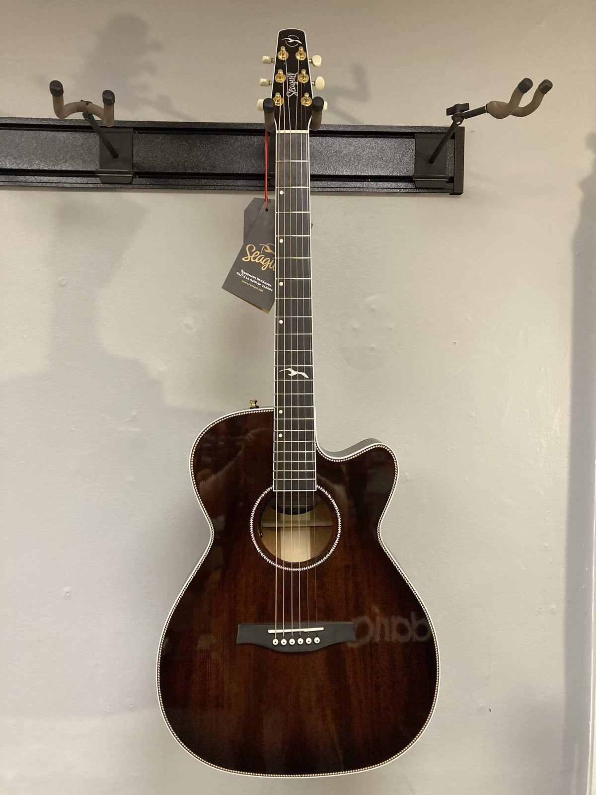 A Seagull Artist Mosaic CH CW Acoustic-Electric Guitar - Bourbon Burst hangs on a wall display. This six-string guitar has gold tuners, a tag on the headstock, a cutaway body, and no visible brand marking on the body.