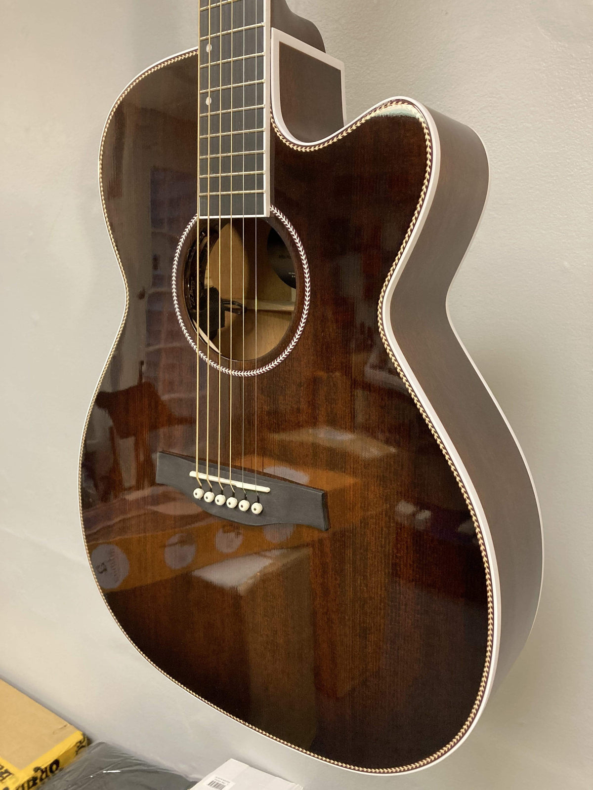 A close-up of the Seagull Artist Mosaic CH CW Acoustic-Electric Guitar - Bourbon Burst (with TRIC Case) shows its glossy finish, white trim, and six strings, with reflections visible as it hangs against a light-colored wall.