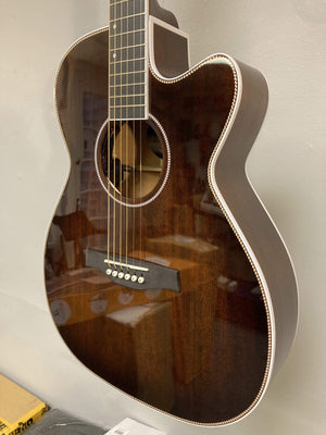 A close-up of the Seagull Artist Mosaic CH CW Acoustic-Electric Guitar - Bourbon Burst (with TRIC Case) shows its glossy finish, white trim, and six strings, with reflections visible as it hangs against a light-colored wall.
