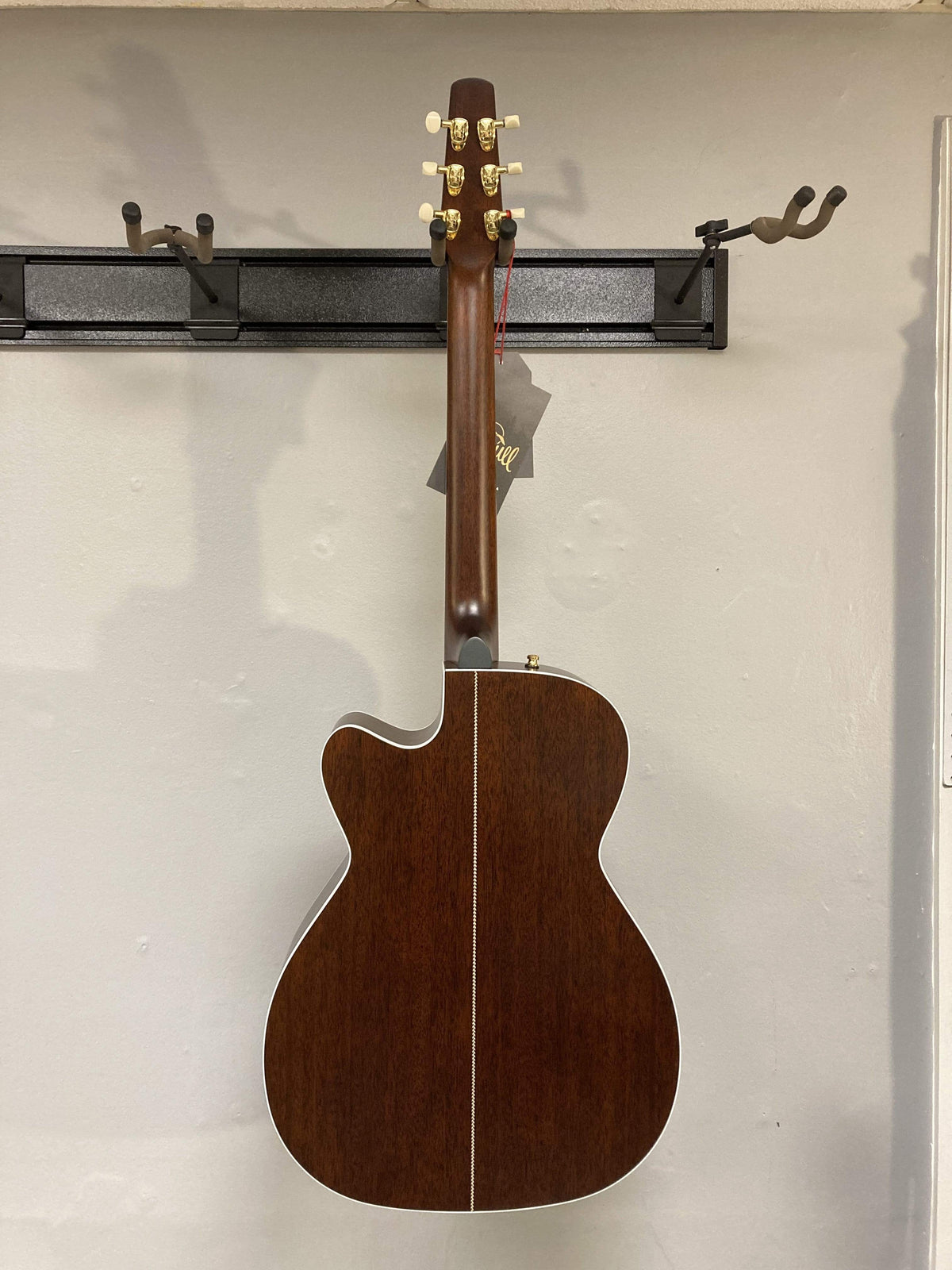 A Seagull Artist Mosaic CH CW Acoustic-Electric Guitar - Bourbon Burst, featuring a natural wood finish, gold tuning pegs, white center stripe on the back, and subtle burst accents, hangs on a wall-mounted rack.