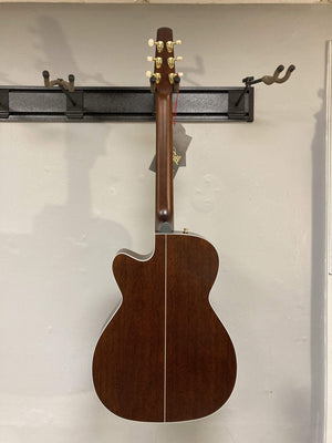 A Seagull Artist Mosaic CH CW Acoustic-Electric Guitar - Bourbon Burst, featuring a natural wood finish, gold tuning pegs, white center stripe on the back, and subtle burst accents, hangs on a wall-mounted rack.