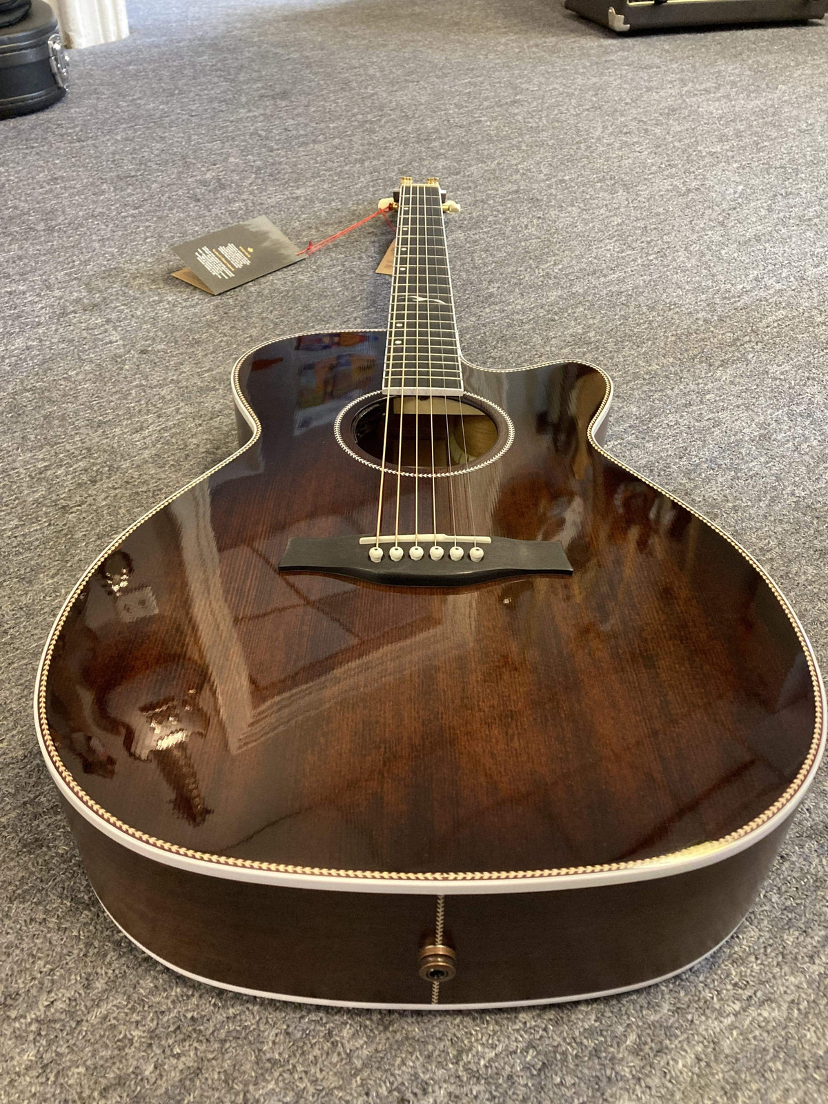 A Seagull Artist Mosaic CH CW Acoustic-Electric Guitar in Bourbon Burst with a cutaway design lies on a gray carpet. A tag hangs from the headstock, and its glossy finish reflects the surroundings. Includes TRIC Case.