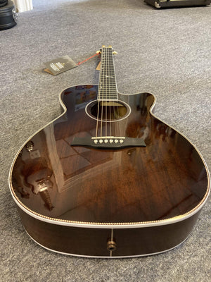 A Seagull Artist Mosaic CH CW Acoustic-Electric Guitar in Bourbon Burst with a cutaway design lies on a gray carpet. A tag hangs from the headstock, and its glossy finish reflects the surroundings. Includes TRIC Case.