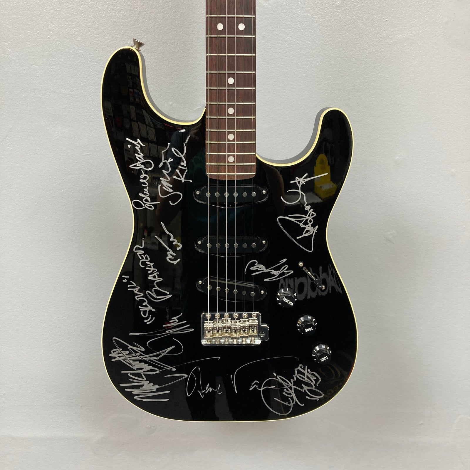 A black electric guitar with a white trim, displayed vertically against a white wall, featuring multiple silver-colored signatures on the body.