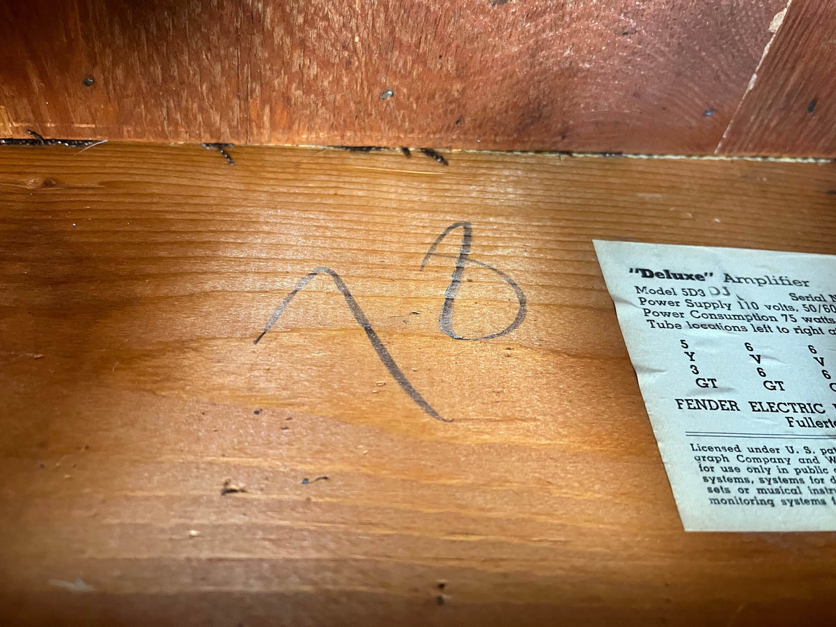 A handwritten "AB" appears on wood near a label about the 1954 Fender Tweed Deluxe Model 5D3 w/Flight Case from Guitars on Main, evoking the classic style of Fender’s vintage tube amplifiers.