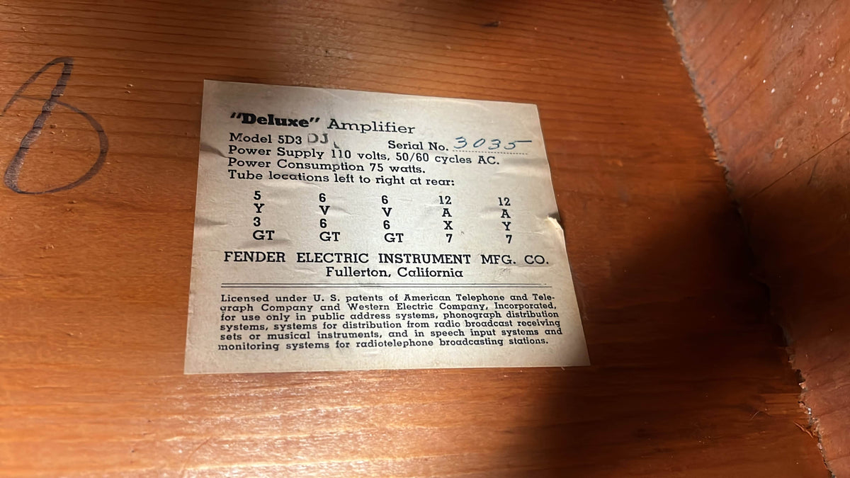 Vintage label on a 1954 Fender Tweed Deluxe Model 5D3 w/Flight Case by Guitars on Main, displaying serial number, power specs, tube layout, and company info atop a wooden surface.