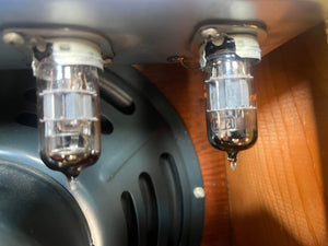 The 1954 Fender Tweed Deluxe Model 5D3 by Guitars on Main features two glass vacuum tubes mounted upside down beside a metal speaker grille and a wooden panel, all protected in its flight case.