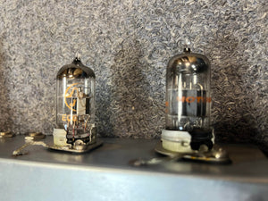 Two glass vacuum tubes are mounted on a metal surface, highlighting the vintage craftsmanship of the 1954 Fender Tweed Deluxe Model 5D3 by Guitars on Main, all set against a textured gray background.