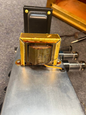 A close-up of the metal transformer on a 1954 Fender Tweed Deluxe Model 5D3 by Guitars on Main, mounted on a chassis beside two vacuum tubes—showcasing authentic hand-wired amp details.
