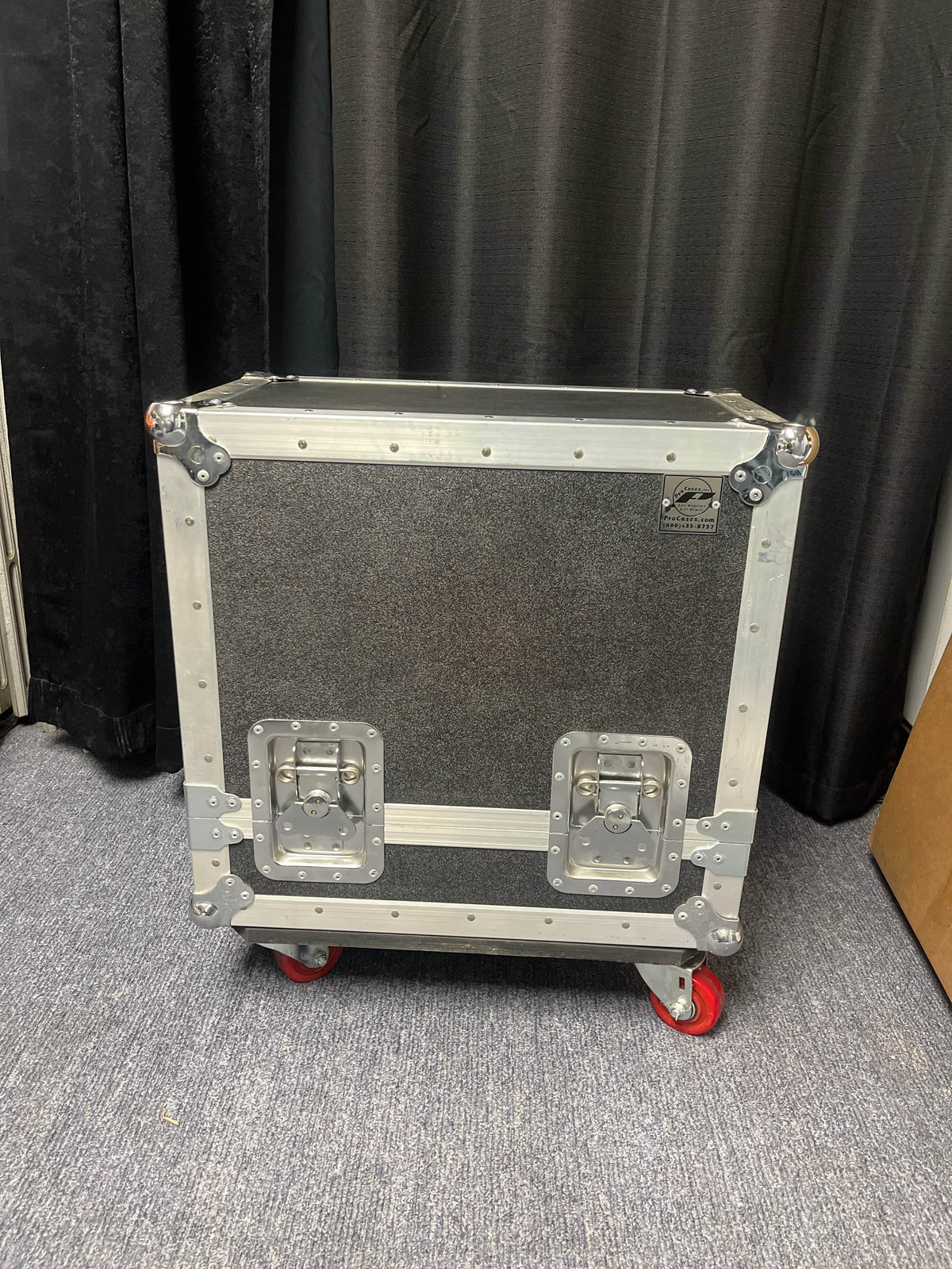 A 1954 Fender Tweed Deluxe Model 5D3 w/Flight Case by Guitars on Main sits on red caster wheels on a carpeted floor before black curtains—the black and silver flight case features metal edges and two latches, ideal for vintage amps.