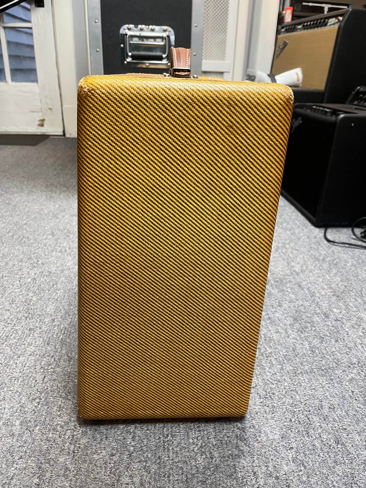 A 1954 Fender Tweed Deluxe Model 5D3 vintage tube amp from Guitars on Main stands upright on a gray carpet, with its flight case and other amps visible in the background.