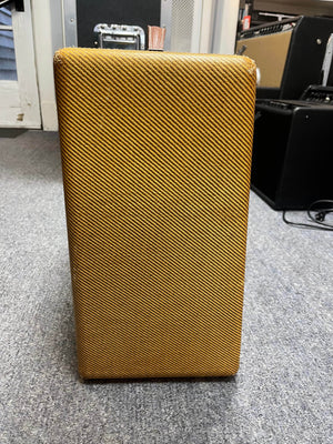 A 1954 Fender Tweed Deluxe Model 5D3 tube amplifier by Guitars on Main stands upright on a gray carpet, with other amps in the background; it includes a flight case.
