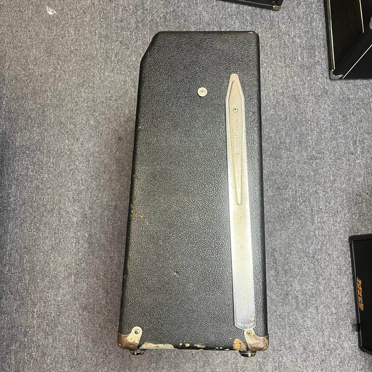 A 1965 Fender Super Reverb - Black Panel from Guitars on Main, featuring a worn vintage look and a long metal handle, lies on its side on a gray carpeted floor. Local pickup available.
