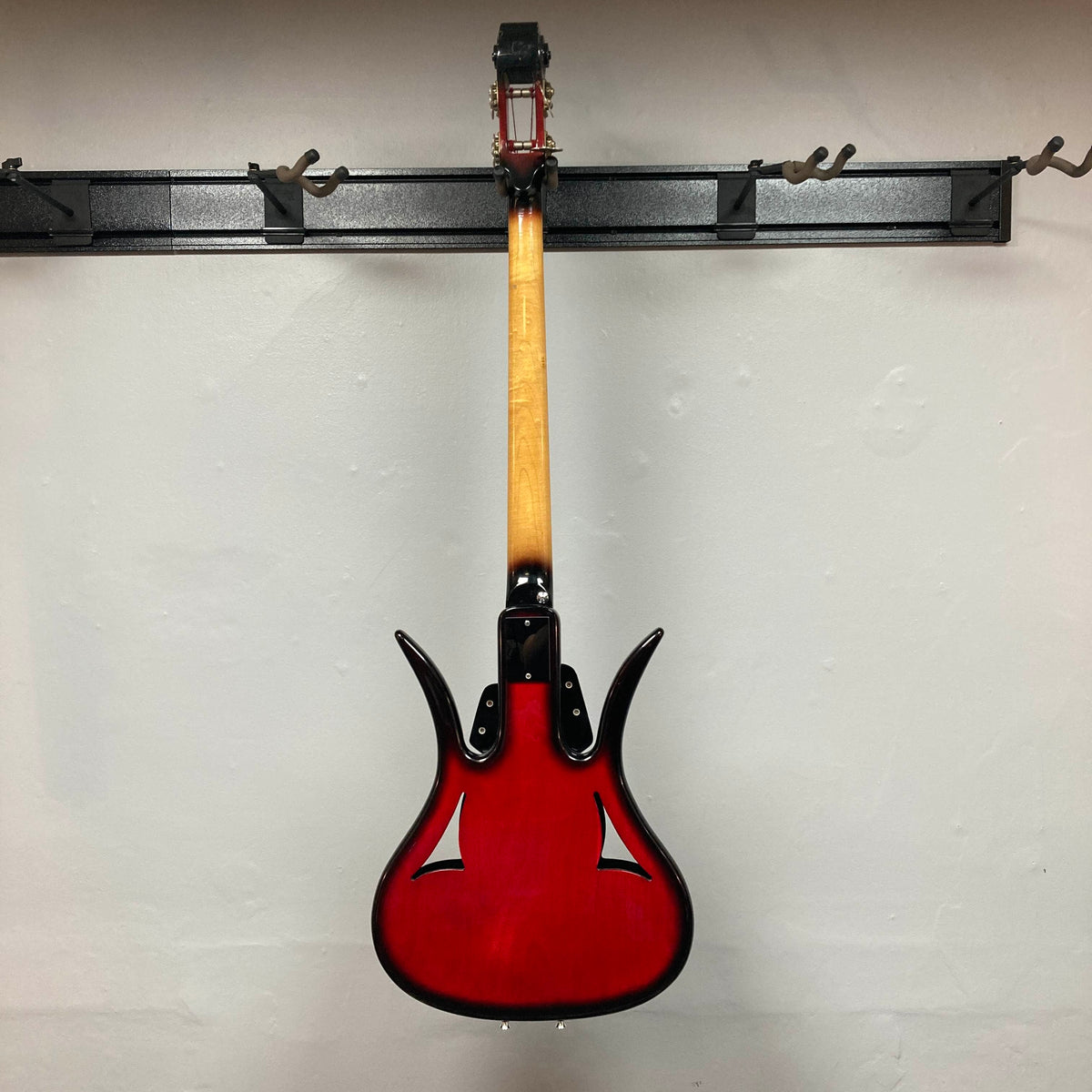 A striking red 1966 Ampeg ASB-1 Devil Bass (Serial #531), featuring a natural wood neck, hangs backward on a black wall-mounted rack to showcase the back of its body and neck, echoing the distinctive vintage design that defines this iconic Ampeg model.