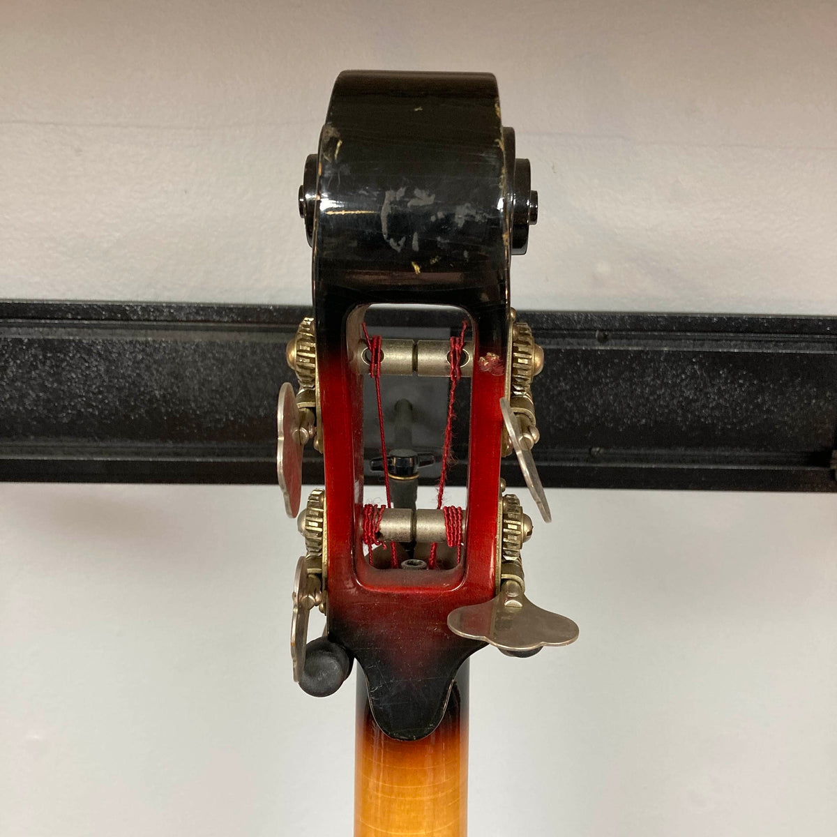 Close-up of the back of a 1966 Ampeg ASB-1 Devil Bass (Serial #531) headstock, highlighting tuning pegs, gears, red string windings, and the wooden neck against a plain wall—capturing the classic details characteristic of this iconic Ampeg instrument.