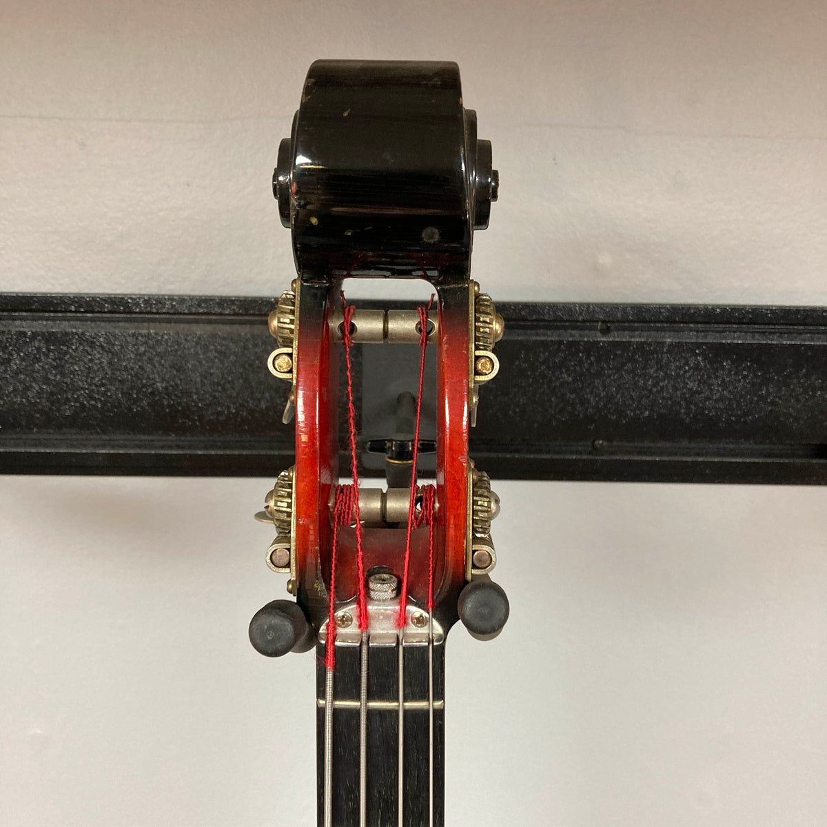Close-up of the top of a string instrument showing tuning pegs, strings, and a black metal support rack against a white background, echoing the distinctive details seen on vintage Ampeg models such as the 1966 Ampeg ASB-1 Devil Bass Serial #531 w/Case.