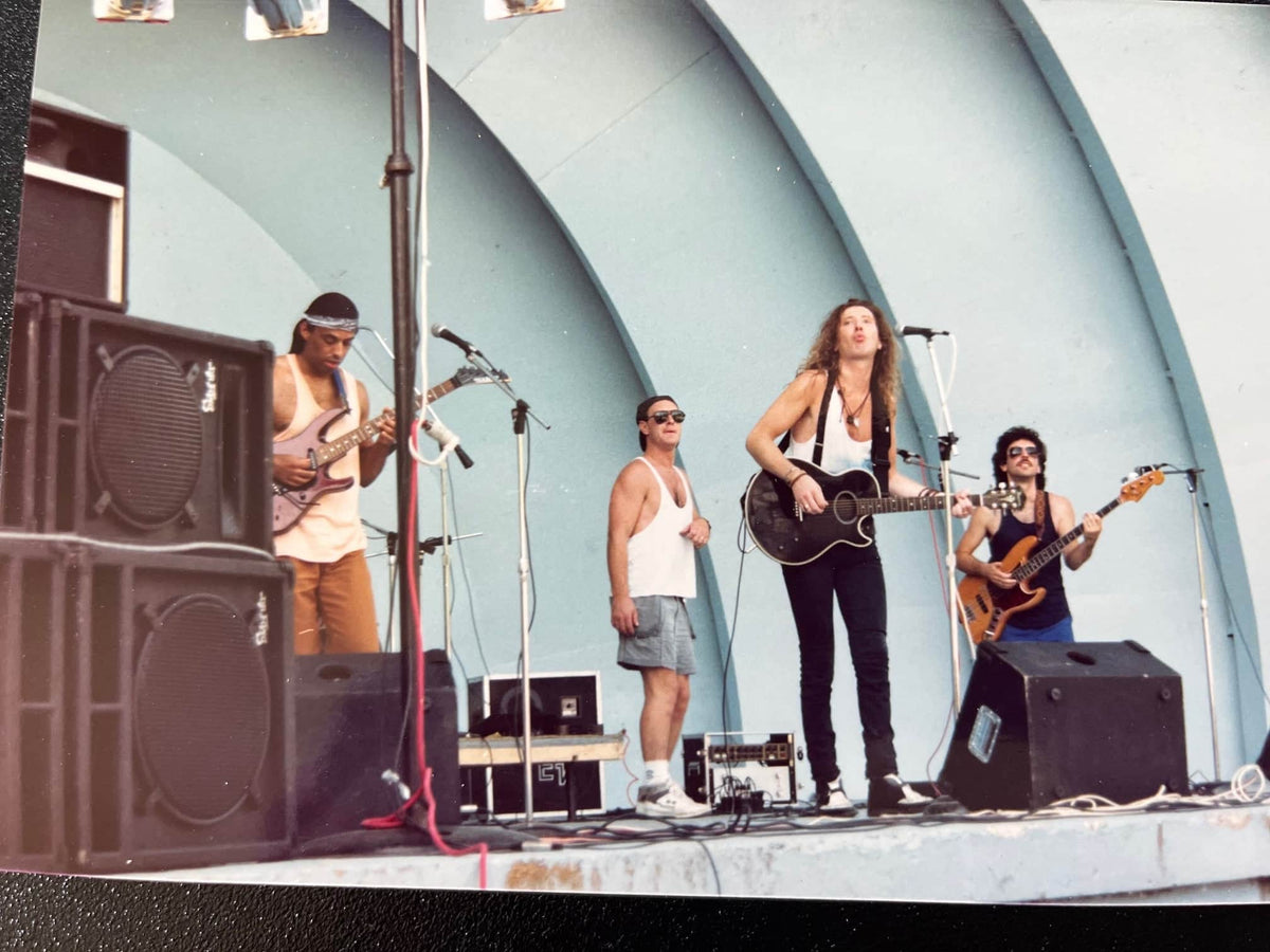 Four musicians perform outdoors—three play guitars, including a Fender 1966 Jazz Bass Natural Refinish—with the bassist using the original pickups. One stands at a mic. Their stage features a curved, shell-like backdrop.