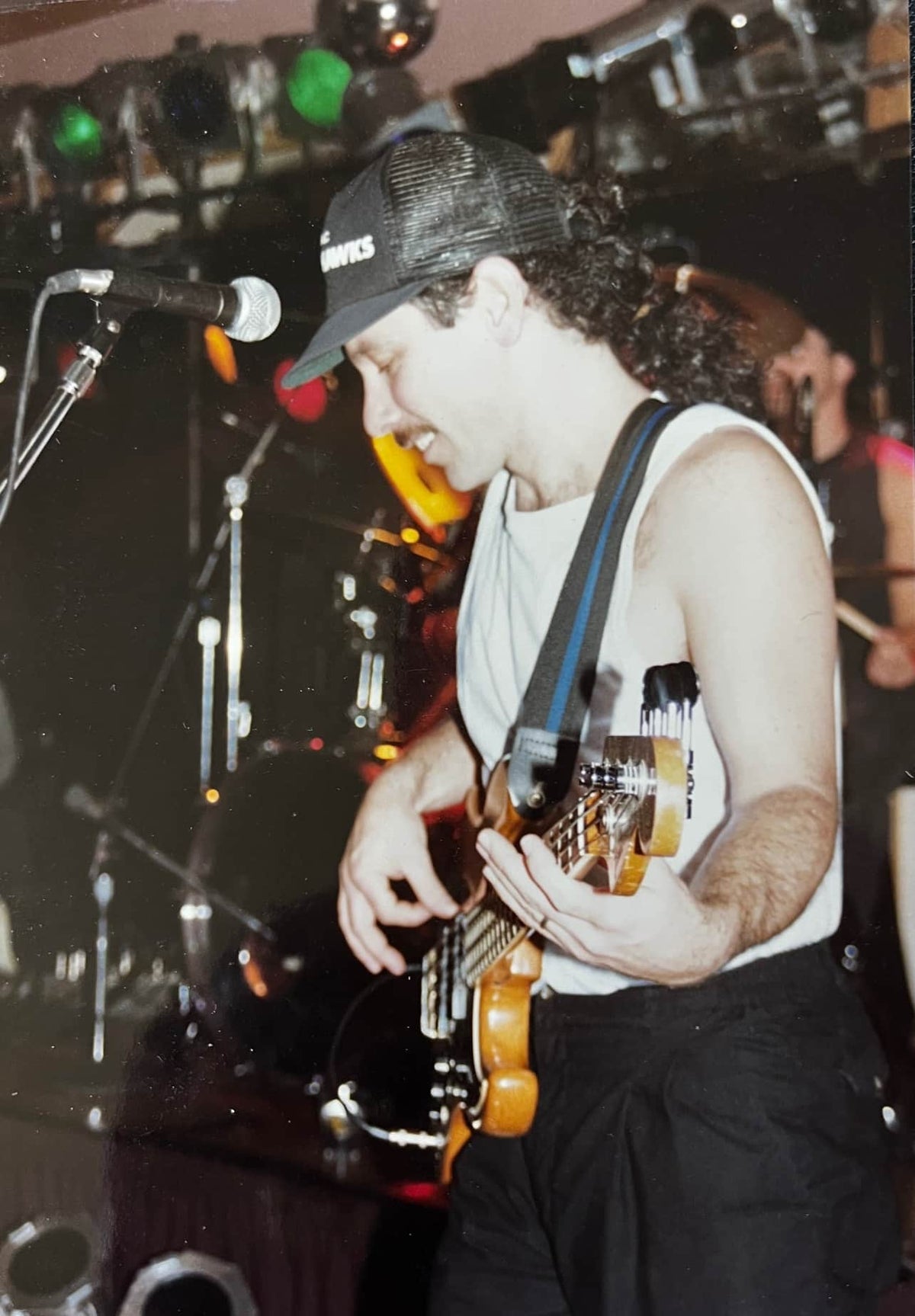 A musician in a cap and sleeveless shirt plays a Fender 1966 Jazz Bass Natural Refinish w/Case on stage, smiling, with drums and stage lights in the background.