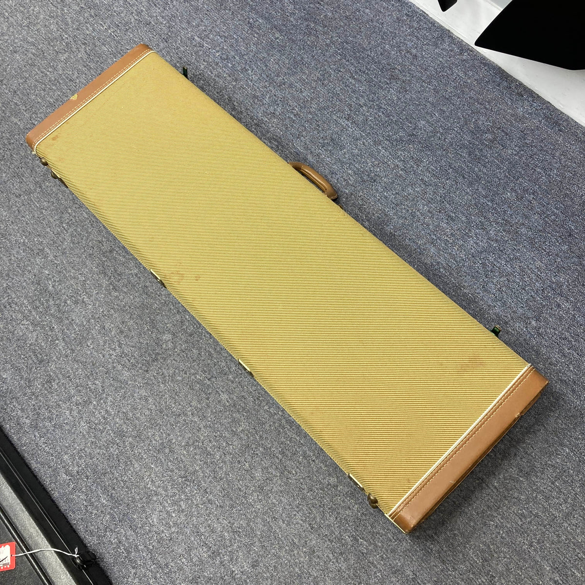 A rectangular, tan and brown hard Fender guitar case holding a 1966 Fender Jazz Bass Natural Refinish rests closed on a gray carpeted floor.