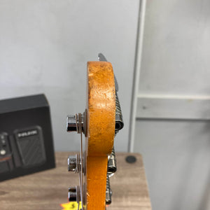 Close-up of a 1966 Fender Jazz Bass Natural Refinish by Fender shows its worn headstock—chips, dents, and tuning pegs visible. In the background: a box and part of a wall, highlighting this vintage bass’s character. Includes case.