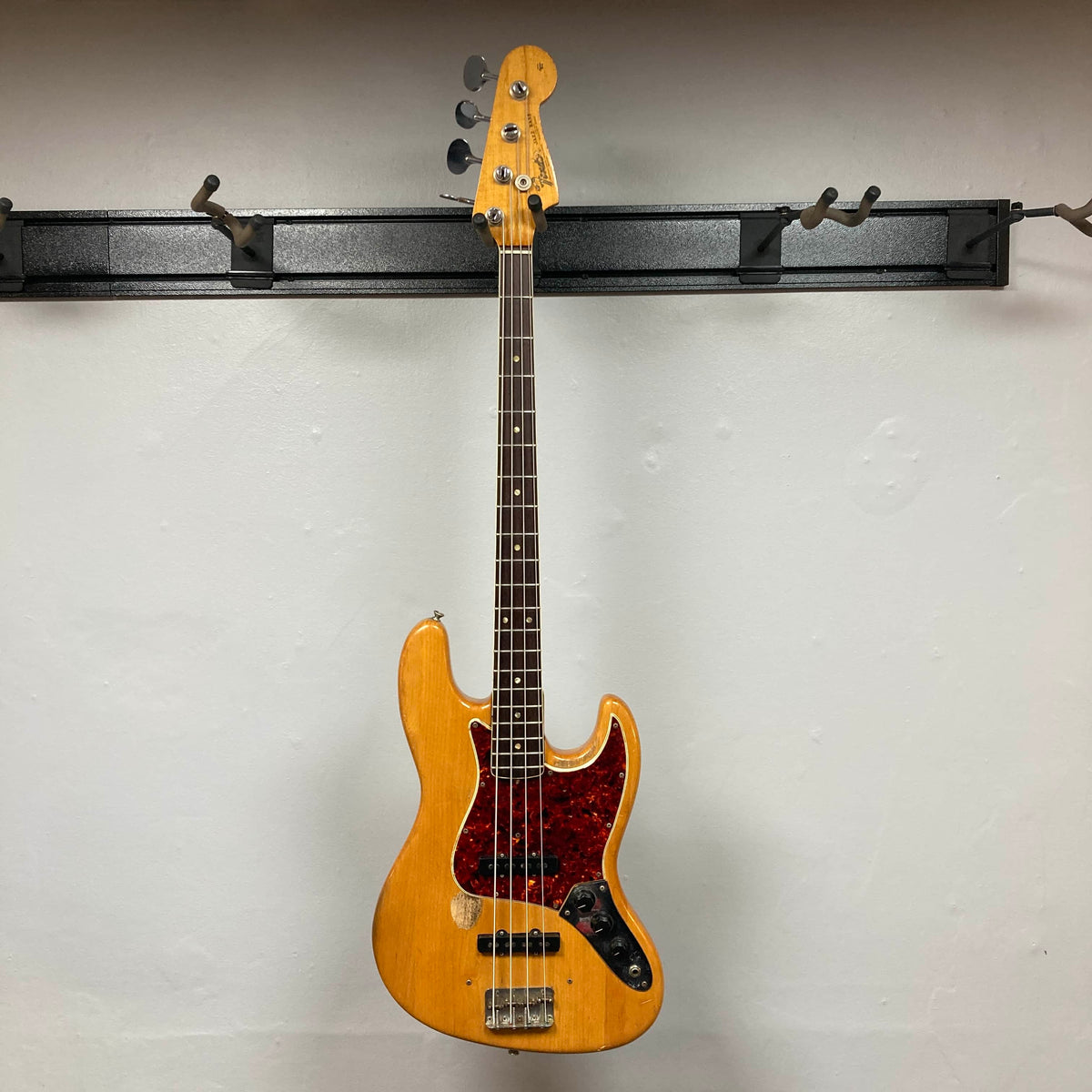 A 1966 Fender Jazz Bass Natural Refinish w/Case by Fender, with original pickups and a tortoiseshell pickguard, hangs on a black wall-mounted rack against a white wall.