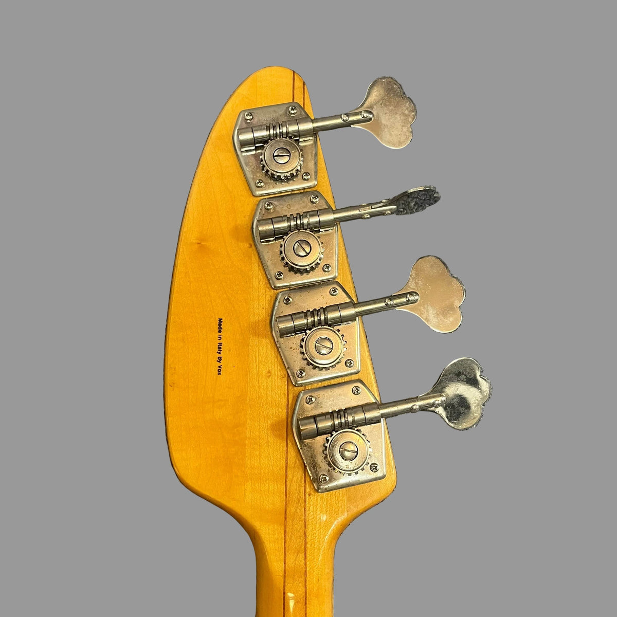 Close-up of the back of a 1967-68 Vox Sidewinder IV Bass (V272) Cherry headstock by Vox, featuring four silver tuning pegs against a plain gray background.